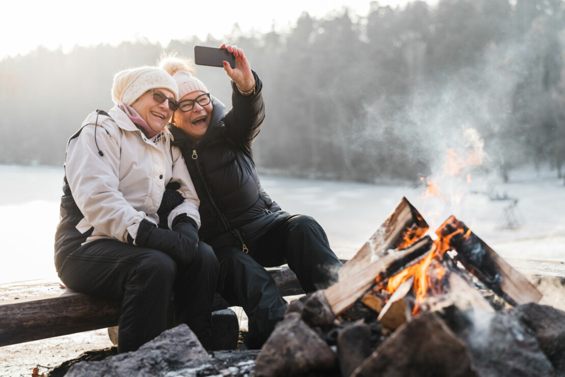 Kaksi nauravaa naista istuu talvella nuotion äärellä ja ottaa selfiekuvan.